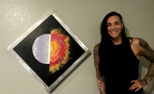 Woman with long dark hair in black sleeveless shirt next to one of her pieces of art, a welded metal frame with a sun in the center, half is polished metal and other other half looks like flames. 