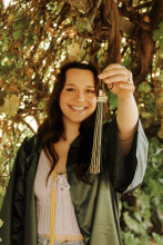 Olivia Bromenschenkel, girl with long dark hair in a green graduation gown holding up a cap tassel showing the 2021 charm.