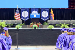 Students sitting in seats facing stage at graduation. Gonfalons displayed on the stage