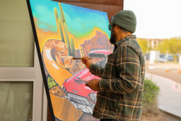 A gentleman wearing a green beenie hat and plaid shirt painting on a canvas