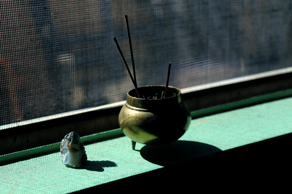 Image of a photograph of a small, brass bowl holding incense sticks, next to a mineral on a green windowsill.