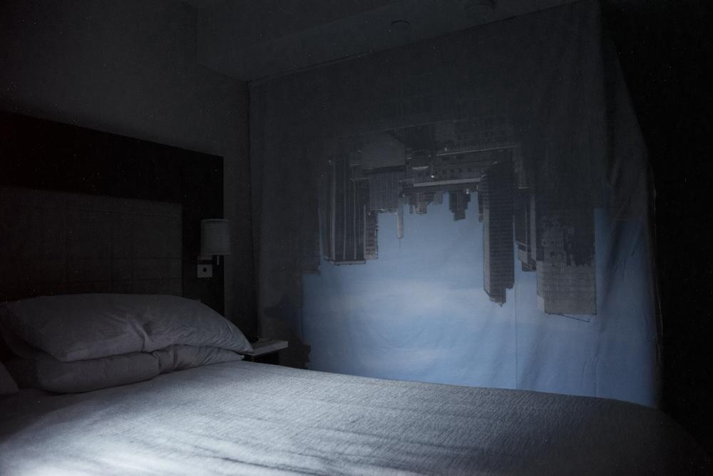Photograph of a bed with a New York skyline upside down on the wall.