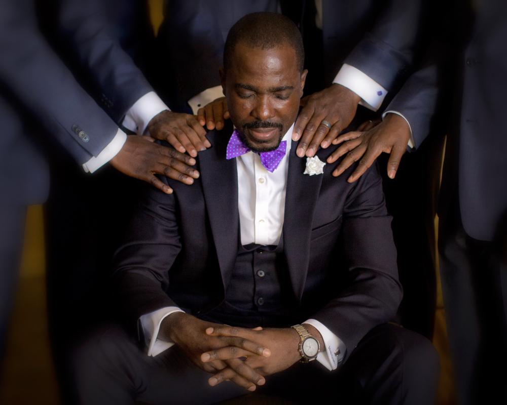 Man in tuxedo in prayer, hands of other people on his shoulders.