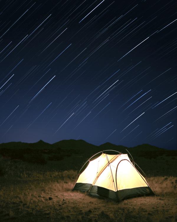 Photograph of an illuminated tent in landscape, under a night sky with star trails held in a long exposure.