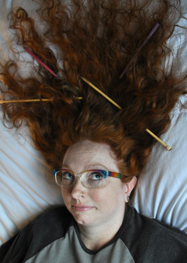 Photograph of the artist with knitting needles and colored pencils in their hair.