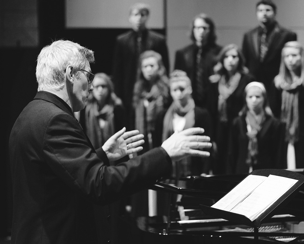 A conductor waves with choir in the background.