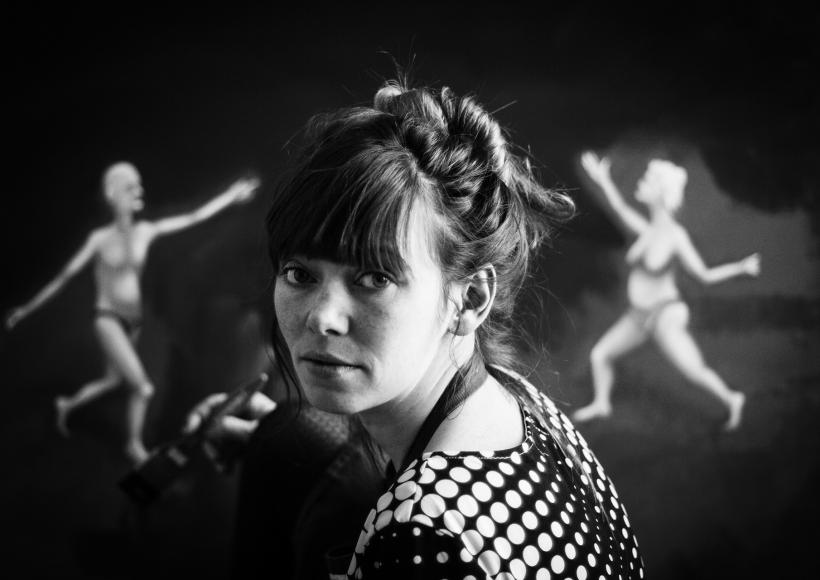 Black and white photograph of girl painting in front of two abstracted figures in the background.