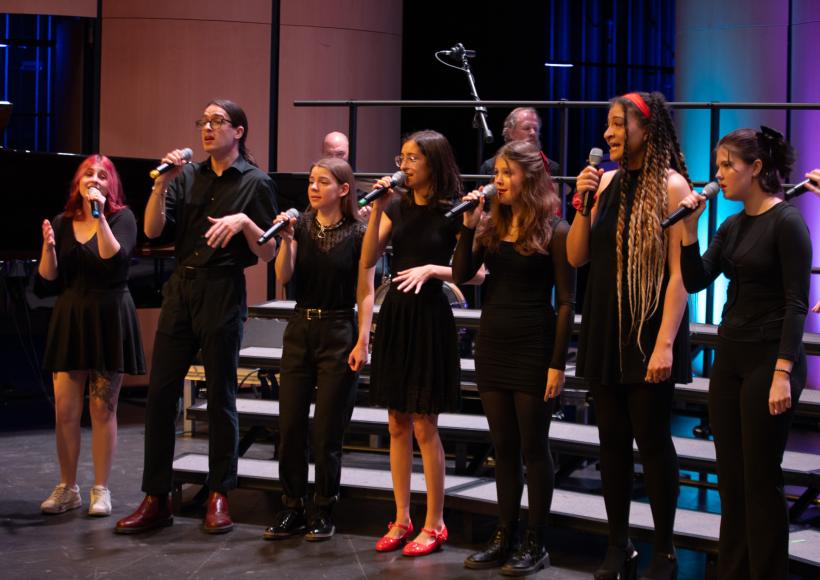 A group of eight performers singing on stage in a line.