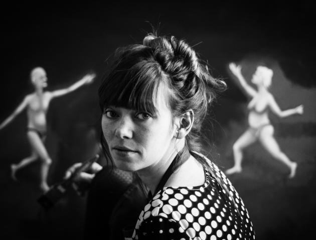 Black and white photograph of girl painting in front of two abstracted figures in the background.