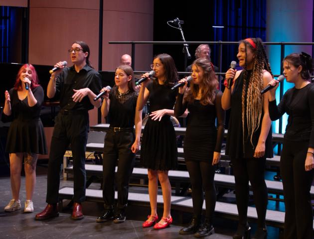 A group of eight performers singing on stage in a line.