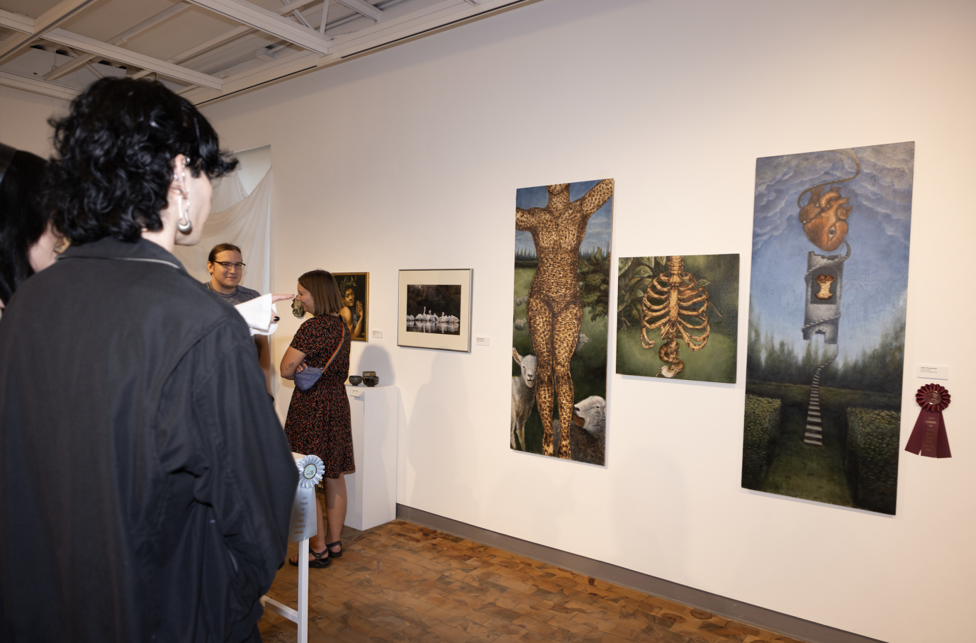 Visitors stand in a bright gallery viewing large surreal paintings, including a leopard-patterned human figure, a ribcage with plants, and a floating heart above a staircase. A red award ribbon is displayed beside one artwork.