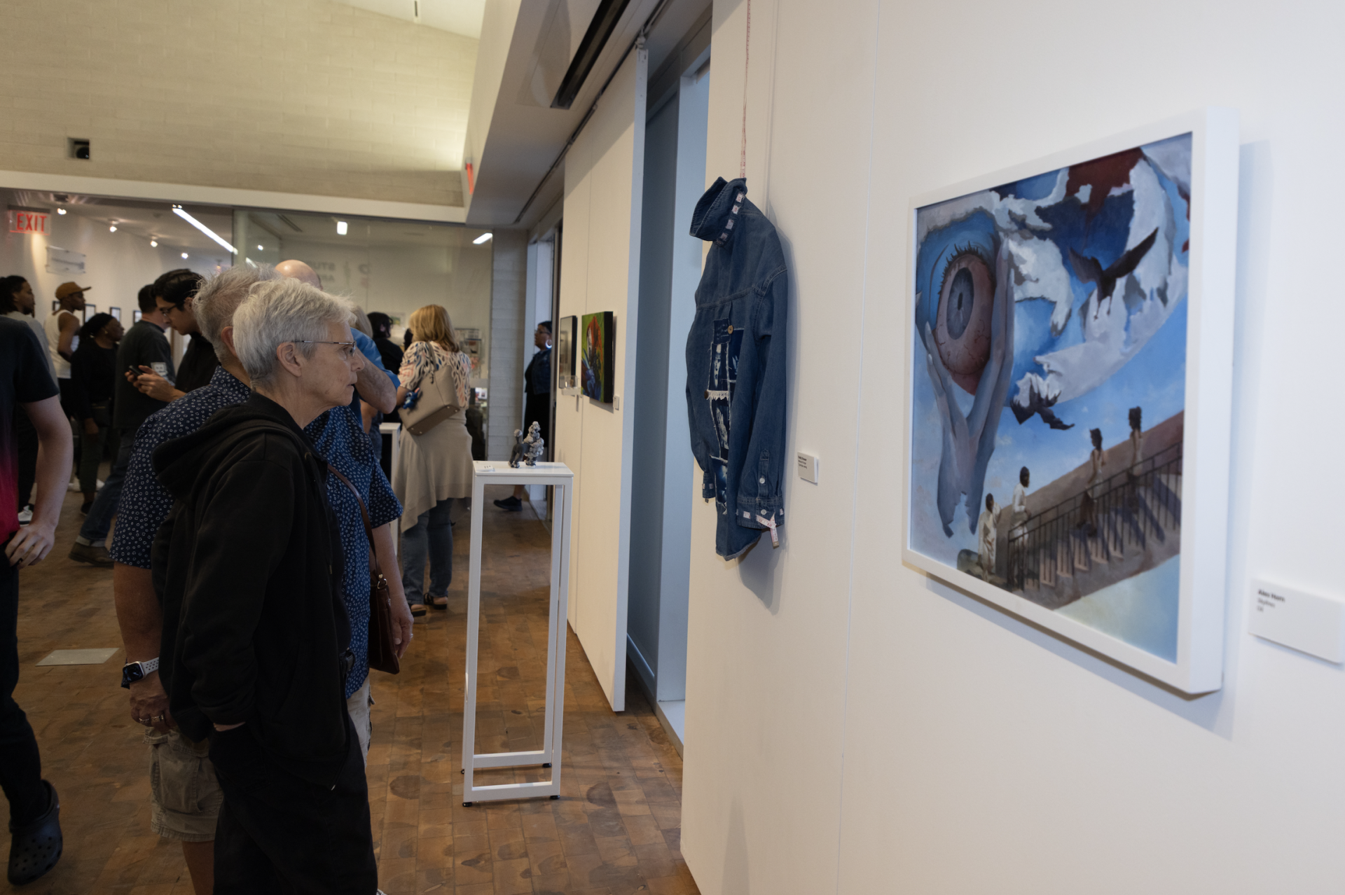 Visitors observe artwork in a gallery. In the foreground, an older woman looks closely at a surreal painting featuring a large eye and flying birds, while a denim jacket is displayed as an art piece nearby.