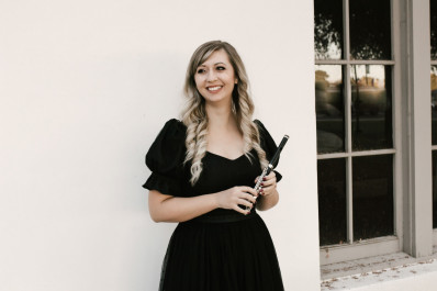 Abby Coffey with long blonde hair curled in a back velvt dress with puffy sleeves holding a wood piccolo standing in front of a light coloured wall near a windowpane