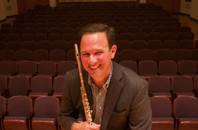 Brian Luce is pictured in a jacket and striped shirt with an all gold flute and concert hall seats in the background behind him