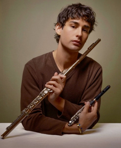 Jarret May wearing a brown shirt leaning on a white table holding a gold flute and a wood piccolo parallel at a 45 degrtee angle