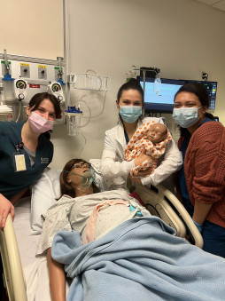 Three nursing students with masks in a hospital simulation room with a woman and baby mannequin.