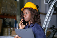 A woman wearing a construction hard hat and holding a clipboard and cell phone
