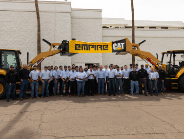 Group of students earning Associate in Applied Science&nbsp;degrees while participating in&nbsp;Empire Southwest’s&nbsp;ThinkBIG apprenticeship program between two large pieces of machinery holding up an Empire CAT banner. 