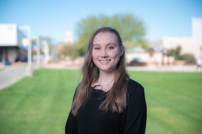 Smiling woman with long sandy brown hair wearing a black top, background is outdoors on an MCC campus with buildings and trees.
