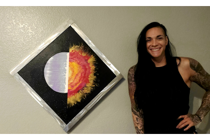 Woman with long dark hair in black sleeveless shirt next to one of her pieces of art, a welded metal frame with a sun in the center, half is polished metal and other other half looks like flames. 