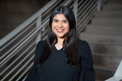 Alejandra Maya -- Portrait of a smiling woman with long dark hair, wearing a black blouse and necklace, standing in front of a stairway with metal railings.