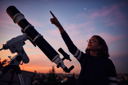 Young girl pointing to the stars standing by a large telescope with evening sky in the background. 