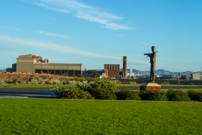 CMC Steel foundry in Mesa, a large beige building with adjacent brick building and stack with a bronze of a steel worker pointing to the building from a grassy area with bushes. Background includes a blue sky with whisky clouds and distant mountains. 