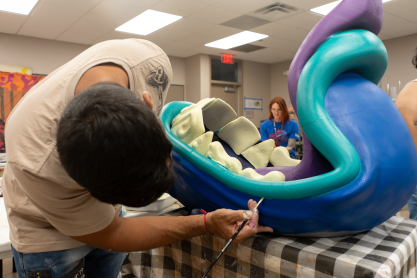 Top of student leaning painting a curved blue structure with large flat teeth. 