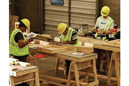 Several people wearing hard hats working with lumber on work tables. 