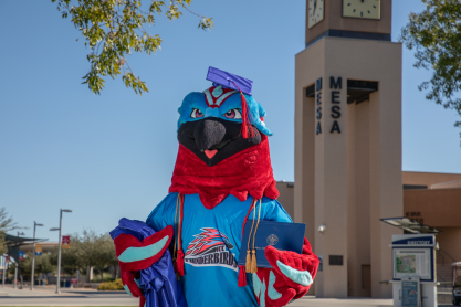 MCC Thunderbird mascot wearing a mortarboard cap holding a graduation gown and diploma cover. The MCC clock tower is in the background. 