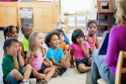 Smiling and laughing children sitting on the floor as a teacher reads a story. 
