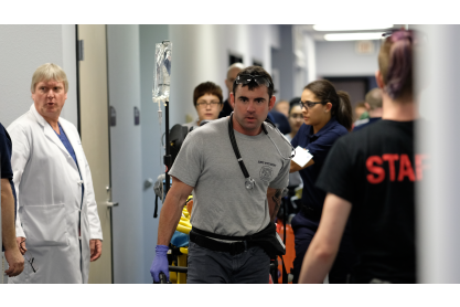 Croweded hallway with an MCC EMT student in a grey t-shirt pulling a gurney, The student has dark hair and a stethoscope around his neck. 