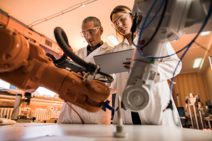 Two people wearing white lab coats and safety glasses operating robotic arms with a tablet. 