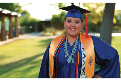 Smiling indigenous female student in blue graduation cap and gown with honor cords and turquoise necklace. 