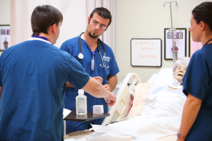 Three students in scrubs around a dummy patient in a hospital bed.