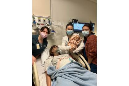 Three MCC nursing students with masks in a simulation delivery room with a mannequin woman and baby.
