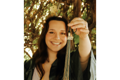 Olivia Bromenschenkel, girl with long dark hair in a green graduation gown holding up a cap tassel showing the 2021 charm.