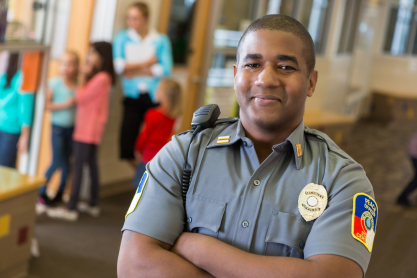 Man in a security guard uniform in the forefront with blurred images of a teacher and young people in the background. 