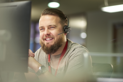 White male with beard wearing a headset, smiling while looking at a computer screen. 
