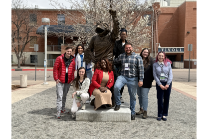 Eight people outside posing with a bronze statue of what appears to be a person in a mascot costume with a bird head. 