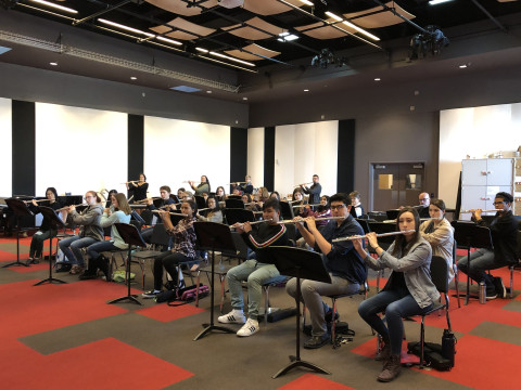 30 high school flutists seated playing group war ups at Flute Regional Etude Day at MCC with red and grey carpet in a liege rehearsal room with white and black acoustic panels