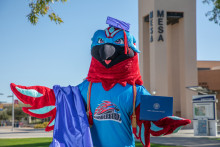 Thor, the MCC Thunderbird mascot-a light blue and red bird, wearing a mortarboard cap holding a diploma cover with a graduation gown draped over one wing. 