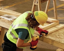 Person wearing a hard hat drilling a piece of lumber on a work table. 