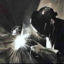 Black and white photo of a person welding. Person is in full gear and the weld is bright sparks. 