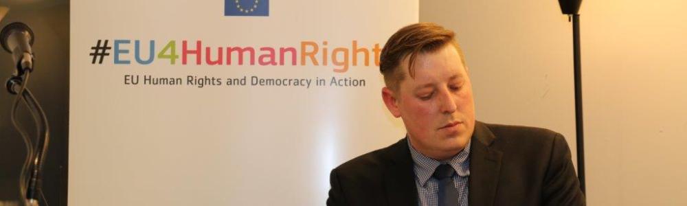 Aaron Holtz in front of a sign for EU Human Rights and Democracy in Action.
