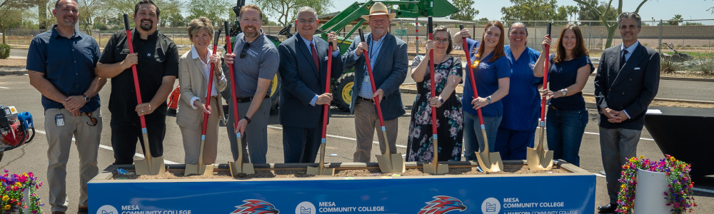 Eleven people standing behind a dirt box holding shovels at a groundbreaking event. 