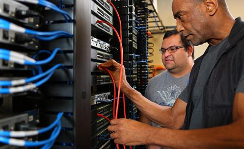 Two people looking at a bank of servers and cables.