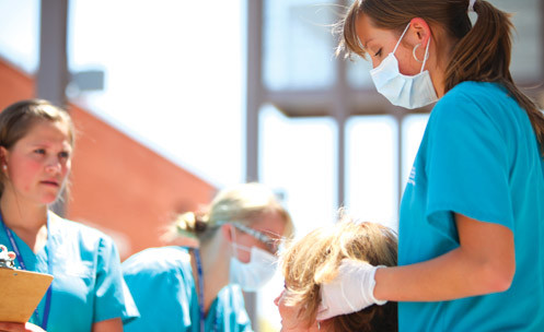 Three students in scrubs, two with masks and one with a clipboard.
