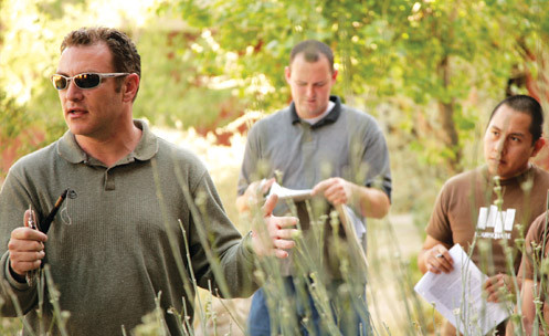 Students watch an instructor outside with shrubs in front.