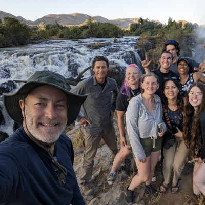 Photo of man with beard and hat next to group of students in front of white water river